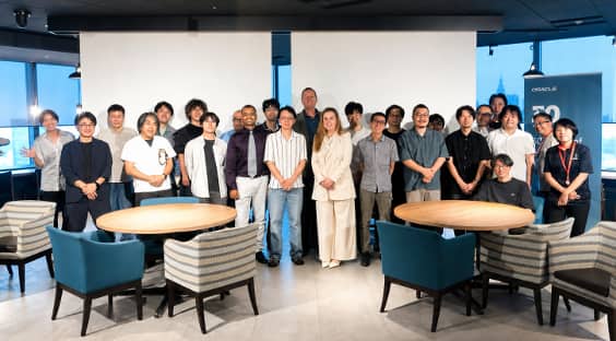 Group of people posing for a photo in a modern office conference area with round tables, large projection screens, and an Oracle sign in the background.