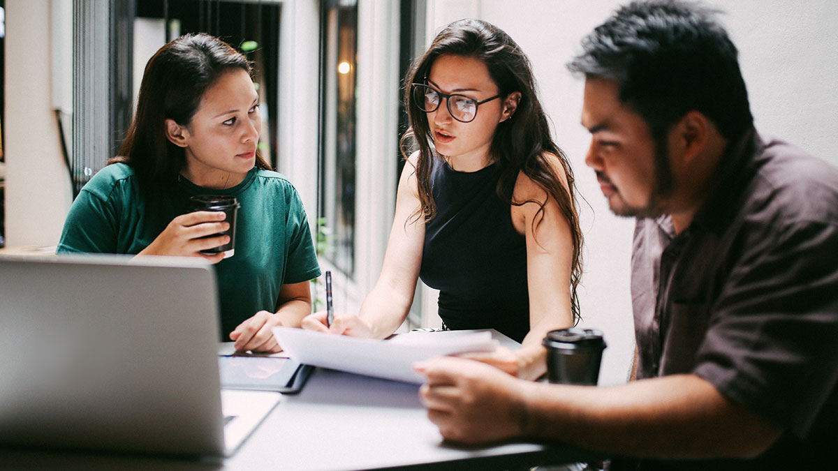Two women and a man sitting at a table reviewing documents with a laptop alongside