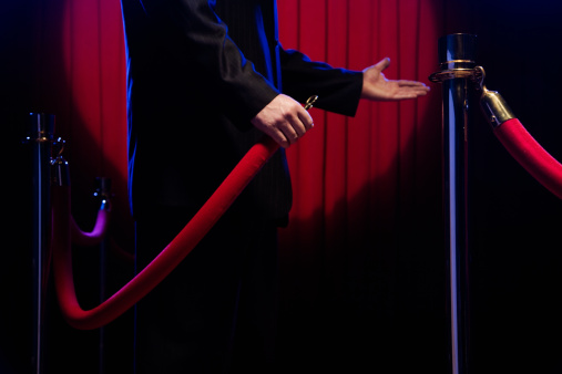 An image of a white man in a blue suit opening a red velvet barricade.